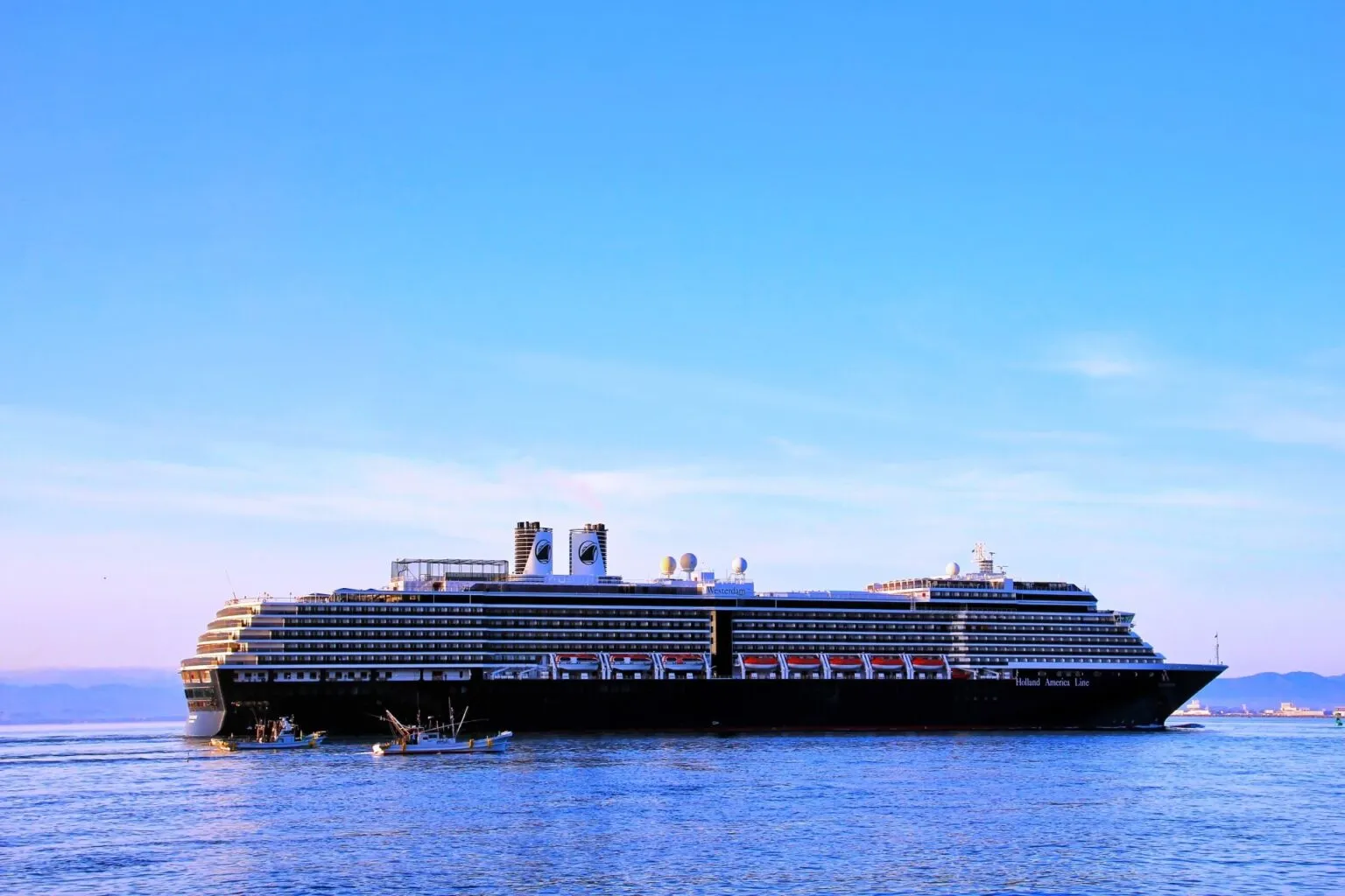 青空の下、海を進むウエステルダム（船体に「Westerdam」の船名）と小さな漁船